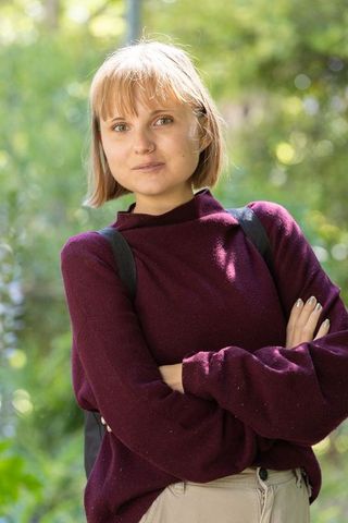 Young woman standing with arms crossed in park wearing maroon sweater and backpack