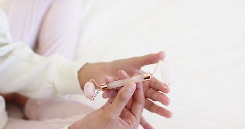 Female hands holding rose quartz facial roller on pale bedding for skincare ritual