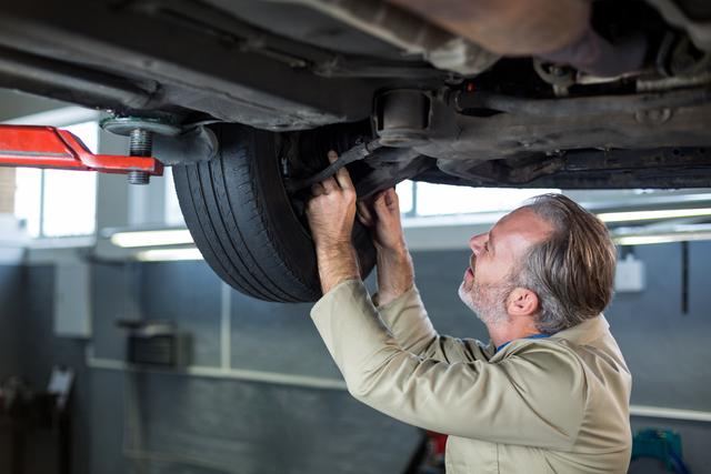 Mechanic is closely examining the undercarriage of a car, ensuring it is properly maintained. Useful for illustrating automotive repair services, maintenance tips, or professional mechanical services. Ideal for websites, advertisements, and articles related to car repair, auto workshops, or vocational training in the automotive industry.