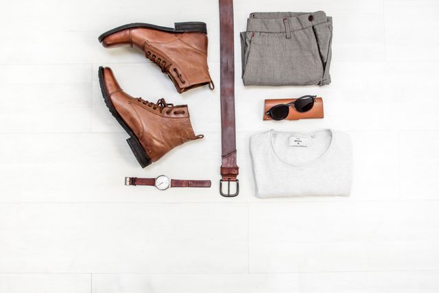 Men's casual outfit displayed in flat lay fashion. Includes leather boots, belt, watch, folded grey t-shirt, brown pants, and sunglasses. Perfect for illustrating men’s fashion, casual wear, styling tips, fashion blogs, social media posts, online stores, and lifestyle magazines.