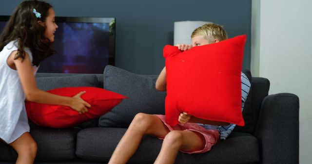 A young girl and boy are having a playful pillow fight in a living room, with copy space. Their cheerful engagement in this common childhood activity creates a lively and joyful atmosphere.