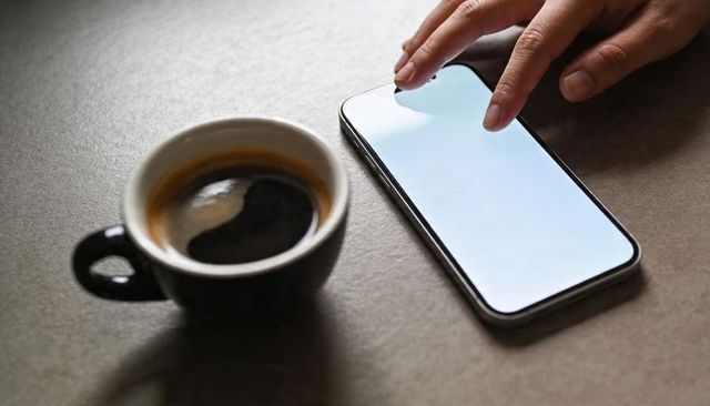 Hand tapping smartphone screen beside espresso cup on coffeehouse table, morning routine