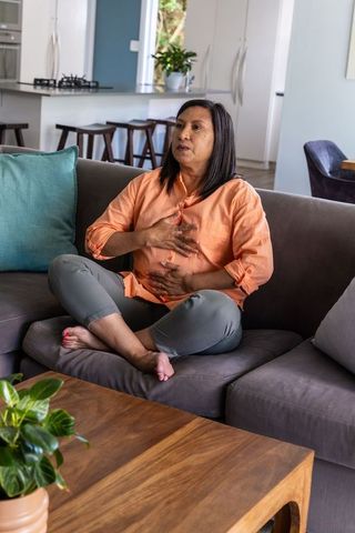 Mature woman experiencing chest and abdominal discomfort on gray sofa in open-plan living room