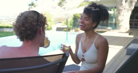 Couple laughing and relaxing by poolside holding citrus drinks on sunlit backyard deck