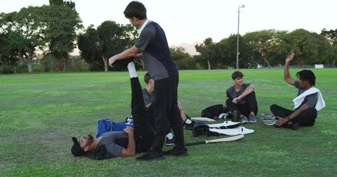 Multiracial cricket teammates stretching and chatting on grass field after practice
