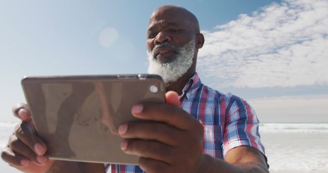 Mature man wearing short-sleeved plaid shirt using tablet on a bright sunny beach day. Perfect for themes like remote work, technology in everyday life, seniors engaging with tech, or relaxation. Great for travel blogs, retirement planning visuals, or lifestyle content focusing on digital devices.