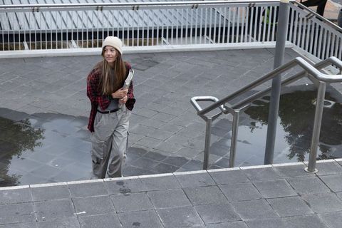 Female student walking up urban stairs holding folders in beanie and plaid shirt, rainy reflections