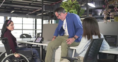 Inclusive team collaborating at desk with wheelchair, tablet and laptop in modern office