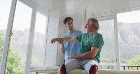 Therapist guiding senior man through shoulder mobility exercise on rehab table with view