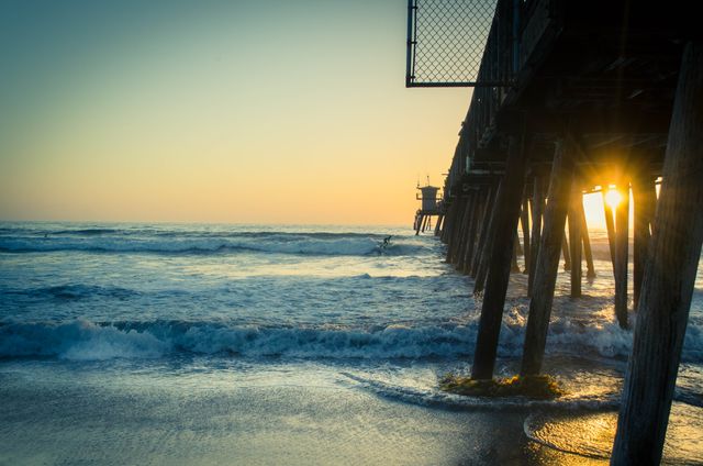 This image captures a tranquil moment at the beach during sunset with waves flowing under a wooden pier. Ideal for promoting beach vacations, coastlines, and serene getaway locations. Great for travel blogs, website banners, and social media posts conveying peace and natural beauty.