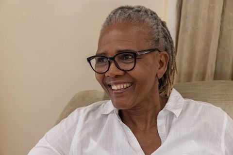 Warm senior african american woman smiling on beige sofa wearing glasses, white shirt