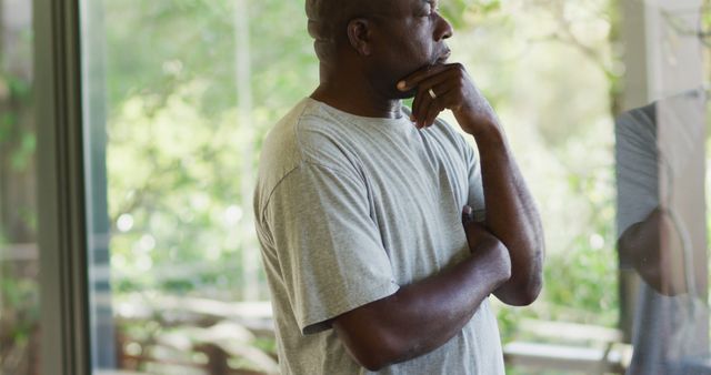 Mature man standing indoors looking pensively out a large window. Natural greenery outside creates a serene and reflective mood. Perfect for conveying themes of introspection, loneliness, contemplation, and quiet moments. Ideal for articles, blogs, and multimedia content focusing on mental health, reflection on life, or personal growth.