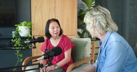 Senior asian woman recording podcast with female host in cozy home studio surrounded by plants