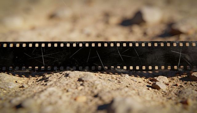Sunlit scratched 35mm film strip lying on sandy ground showing sprocket holes and grain