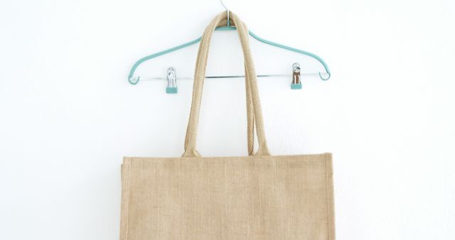 Shot of a reusable jute bag hanging on a clear wire hanger against a plain white wall. Ideal for themes related to sustainability, environmental consciousness, minimalism, and zero waste. Can be used in articles, banners, or social media posts promoting eco-friendly products or lifestyle choices.