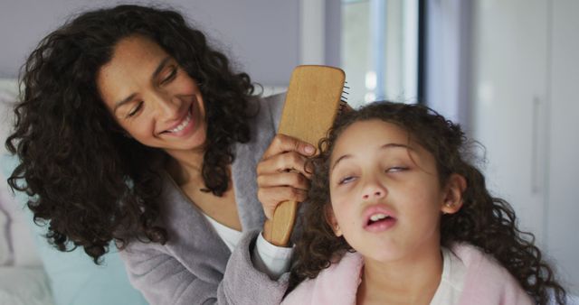 Mother combs her daughter's curly hair with a wooden brush, in an indoor setting, possibly during a morning routine. The scene highlights the close bond and loving care between parent and child. This image is ideal for use in articles about parenting, haircare tips, family bonding, or advertising family-oriented products.
