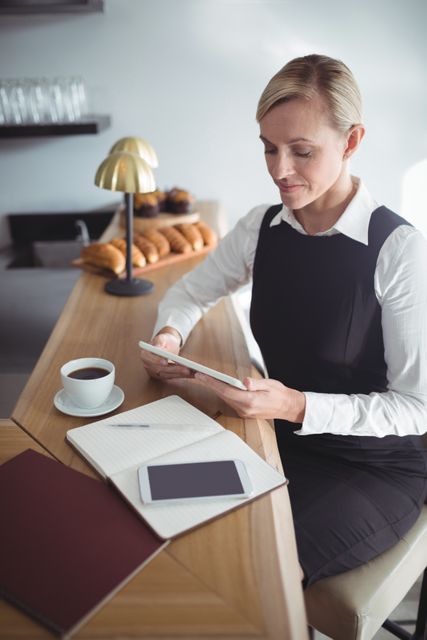 Waitress in uniform using digital tablet at restaurant counter, with coffee and notebook nearby. Ideal for themes related to hospitality, modern technology in service industry, professional work environment, and restaurant management.