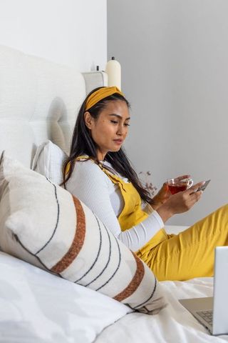 Asian woman remote working on bed in mustard overalls, holding tea and smartphone
