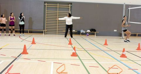 Coach directing diverse women's volleyball agility drill with cones, rings and net