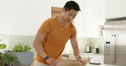 Asian man unpacking groceries on modern bright kitchen island in casual orange t-shirt