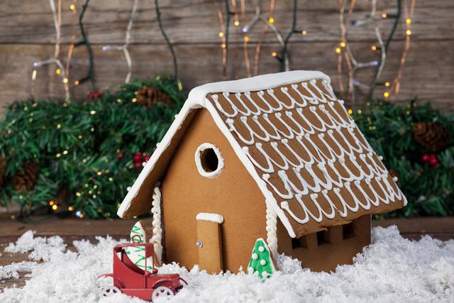 Gingerbread house decorated with icing, surrounded by artificial snow and a toy car, set against a backdrop of Christmas lights and greenery. Ideal for holiday-themed promotions, Christmas cards, festive advertisements, and seasonal blog posts.