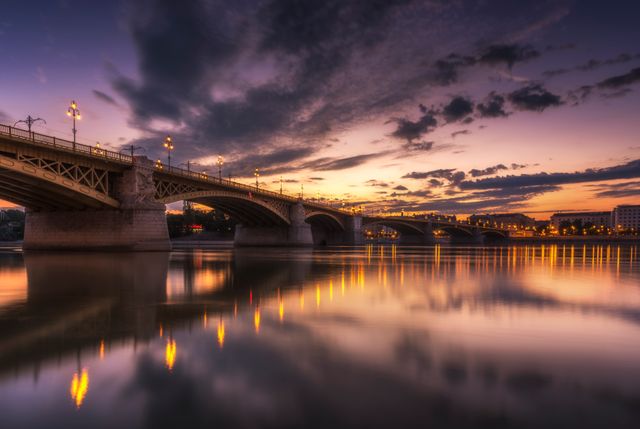 Beautiful scenic view of a city bridge illuminated by streetlights at sunset with vibrant colors reflecting on the calm river. Ideal for travel websites, postcards, or inspirational backgrounds showcasing urban landscapes and serenity.