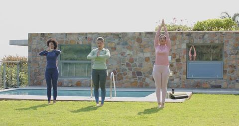 Multicultural women practicing morning yoga on lawn by pool for wellness and mindfulness