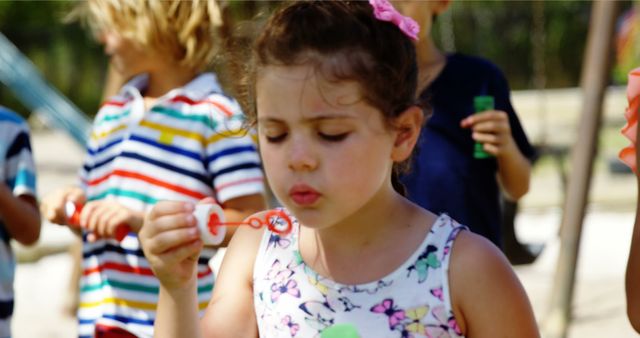 A young girl wears a floral dress while blowing bubbles outdoors at a playground, surrounded by other children. Perfect for use in advertisements, educational materials, parenting blogs, and content focusing on children's activities, outdoor fun, and social interactions.