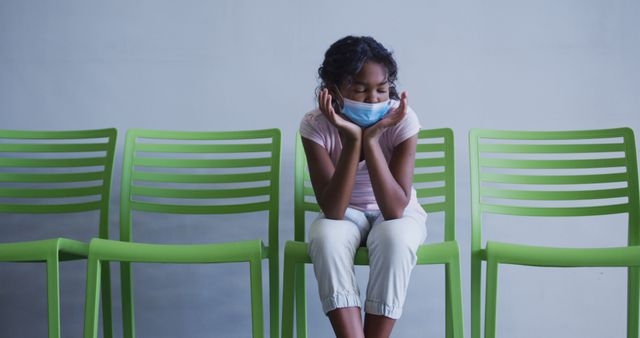 African american girl lowering her face mask while sitting on a chair at hospital. medical healthcare during coronavirus covid 19 pandemic concept