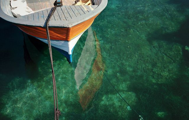 Traditional wooden boat moored in calm, clear blue water showing a beautiful reflection on the surface. Uses include travel blogs, nautical-themed websites, and magazines emphasizing nature, relaxation, and marine activities.