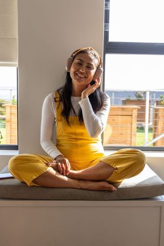 Sunlit asian woman sitting on window seat in yellow overalls listening on pink headphones