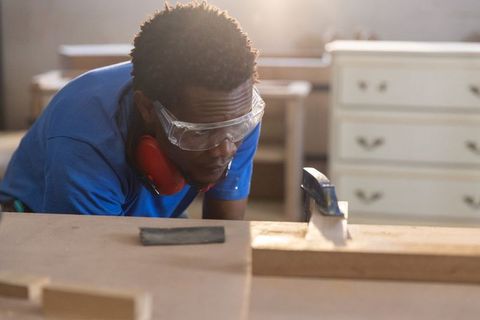 African american carpenter inspecting clamped wood plank in workshop wearing safety goggles
