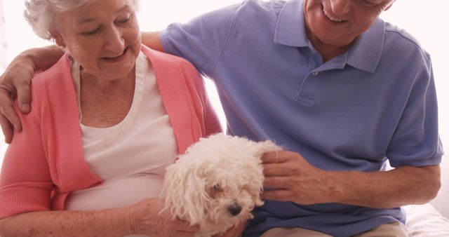 This image depicts a happy older couple smiling and affectionately petting a small, fluffy white dog at home. It conveys a sense of love, companionship, and bond between the seniors and their pet. The image is suitable for use in articles or advertisements about senior living, pet therapy, retirement, elderly lifestyle, family bonding, and home care for seniors.