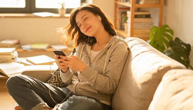 Young woman relaxing on sunlit sofa listening to music with smartphone and wired earbuds