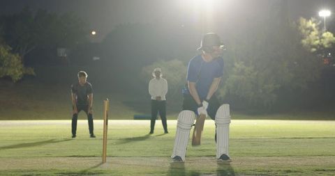Teenage batsman batting under floodlights on night cricket field with helmet and pads