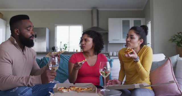 Family of three sitting on a couch and enjoying pizza with wine in a cozy living room. They appear to be engaging in conversation and spending quality time together. Ideal for themes related to family bonding, home dining, casual gatherings, and relaxed moments.