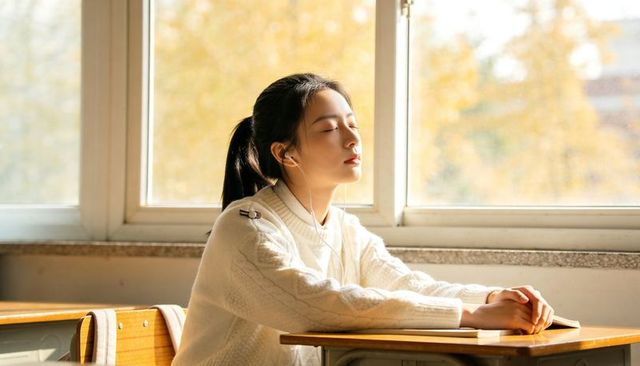 Teen student relaxing at desk with earbuds in sunlit autumn classroom wearing white sweater
