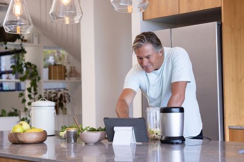 Mature man preparing green smoothie and checking tablet in modern sunlit kitchen