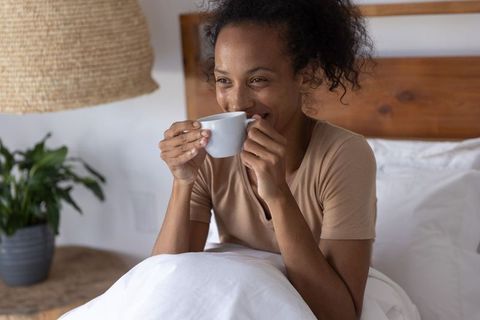 African american woman enjoying morning coffee in bed, smiling, cozy natural light
