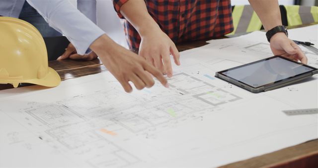 Architects are collaborating on building plans, one person holding a tablet and both pointing at blueprints. Suitable for illustrating concepts of teamwork, construction projects, architectural planning, and engineering. Useful for articles, websites, educational materials, and presentations on architecture and construction management.