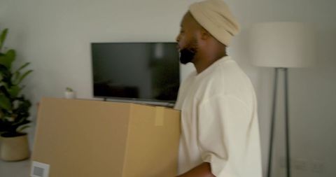 Young man carrying cardboard box in modern minimalist living room with tv and floor lamp