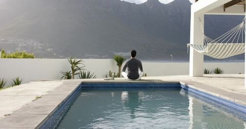 Indian man meditating on poolside mat facing ocean bay with hammock and mountain view