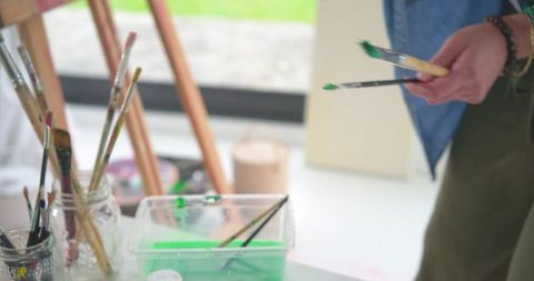 Artist Selecting Paintbrushes at Sunlit Studio Table with Green Paint Tub and Jars