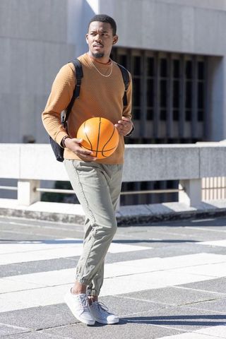African american man walking with basketball on sunlit urban plaza wearing brown sweater
