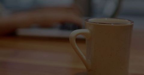 Cozy workspace with speckled ceramic mug sitting near blurred laptop keyboard and hands
