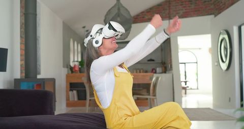 Mature woman enjoying vr headset at home wearing yellow overalls reaching forward