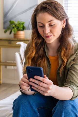 Young woman scrolling smartphone on sunlit sofa holding navy phone for lifestyle content