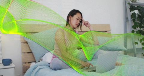 Korean woman working remotely on laptop in bed with neon data wave overlay