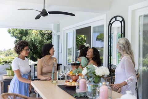 Diverse women hugging and celebrating on sunlit porch with cupcakes, champagne, flowers