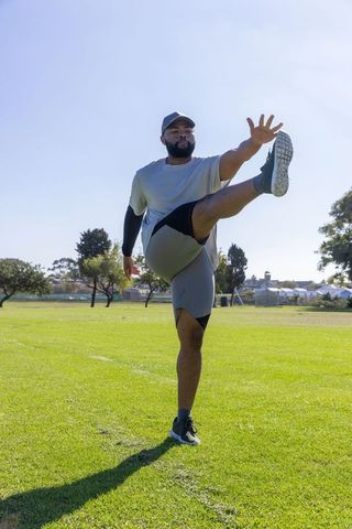 African american man performing high kick while balancing on one leg in sunny park workout